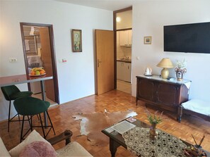 Una sala de estar con un sofá, una mesa de comedor con sillas, una cocina al fondo, un televisor de pantalla plana montado en la pared y una pequeña mesa con un florero y una lámpara.