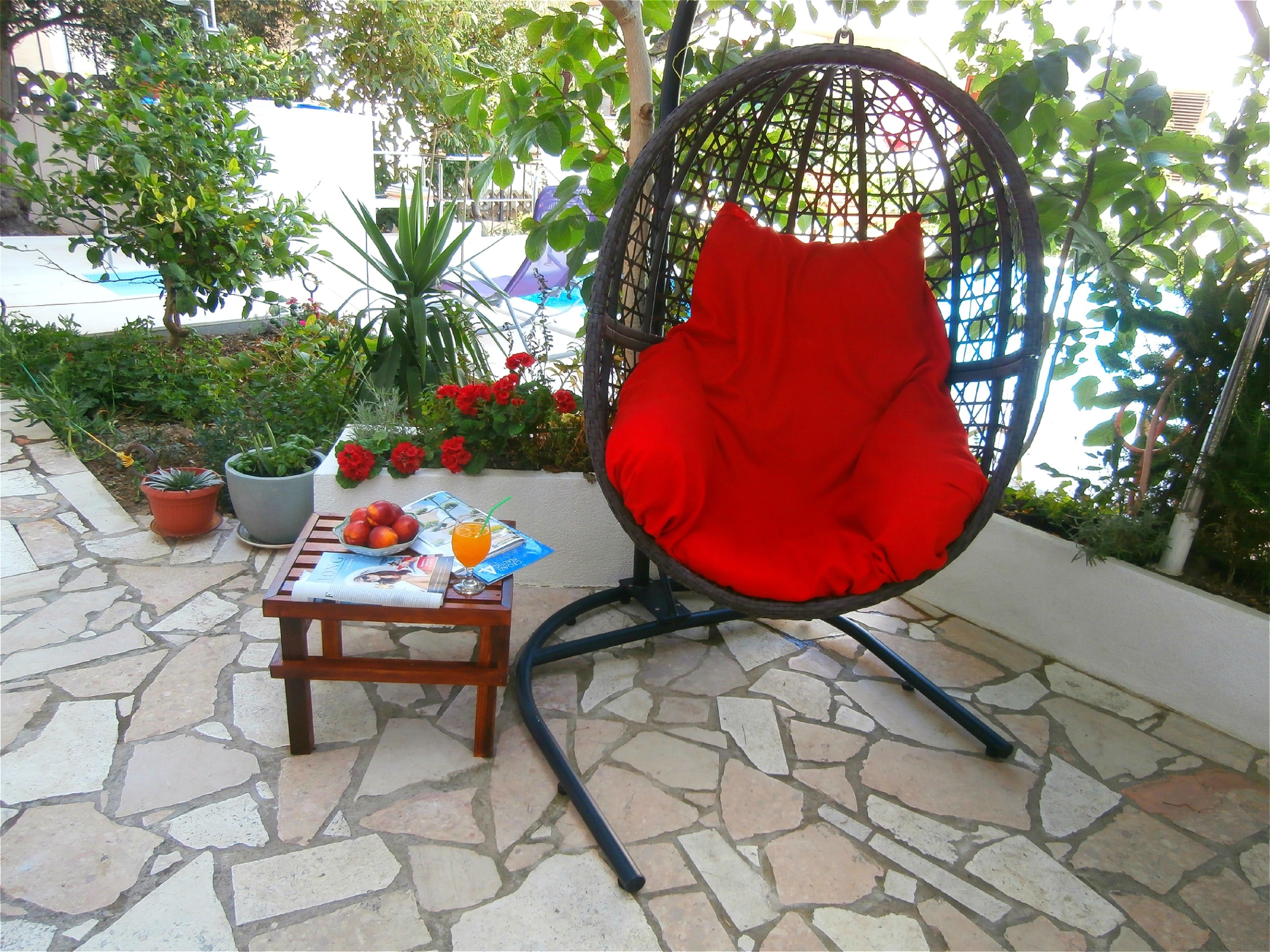 A wicker chair with a red cushion, a small table with a newspaper, fruit, and a drink, and potted plants in a garden setting.