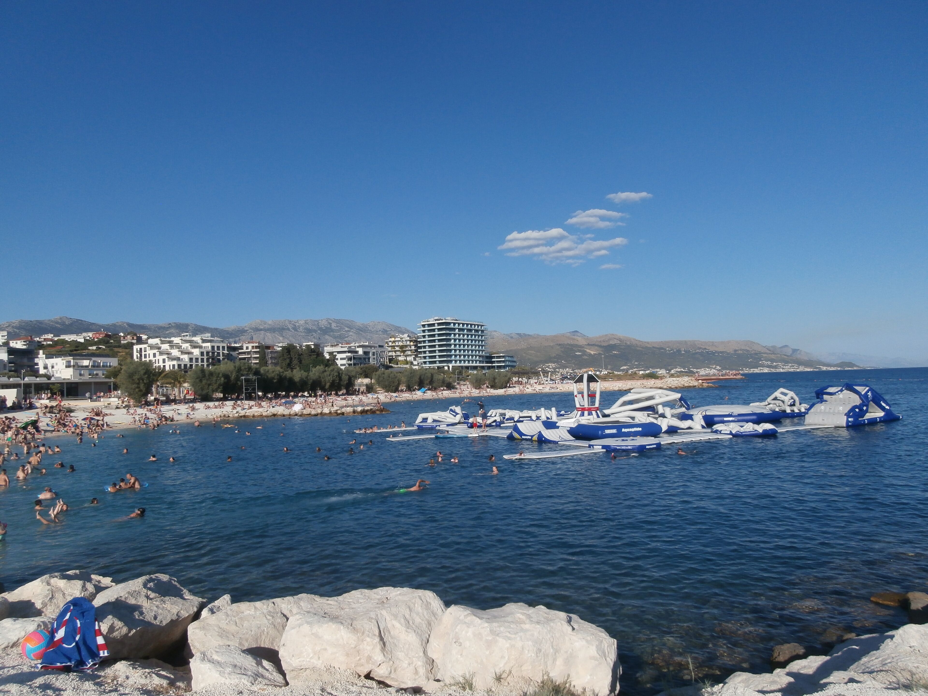 A beach with people swimming and a large inflatable structure in the water. There are buildings in the background.