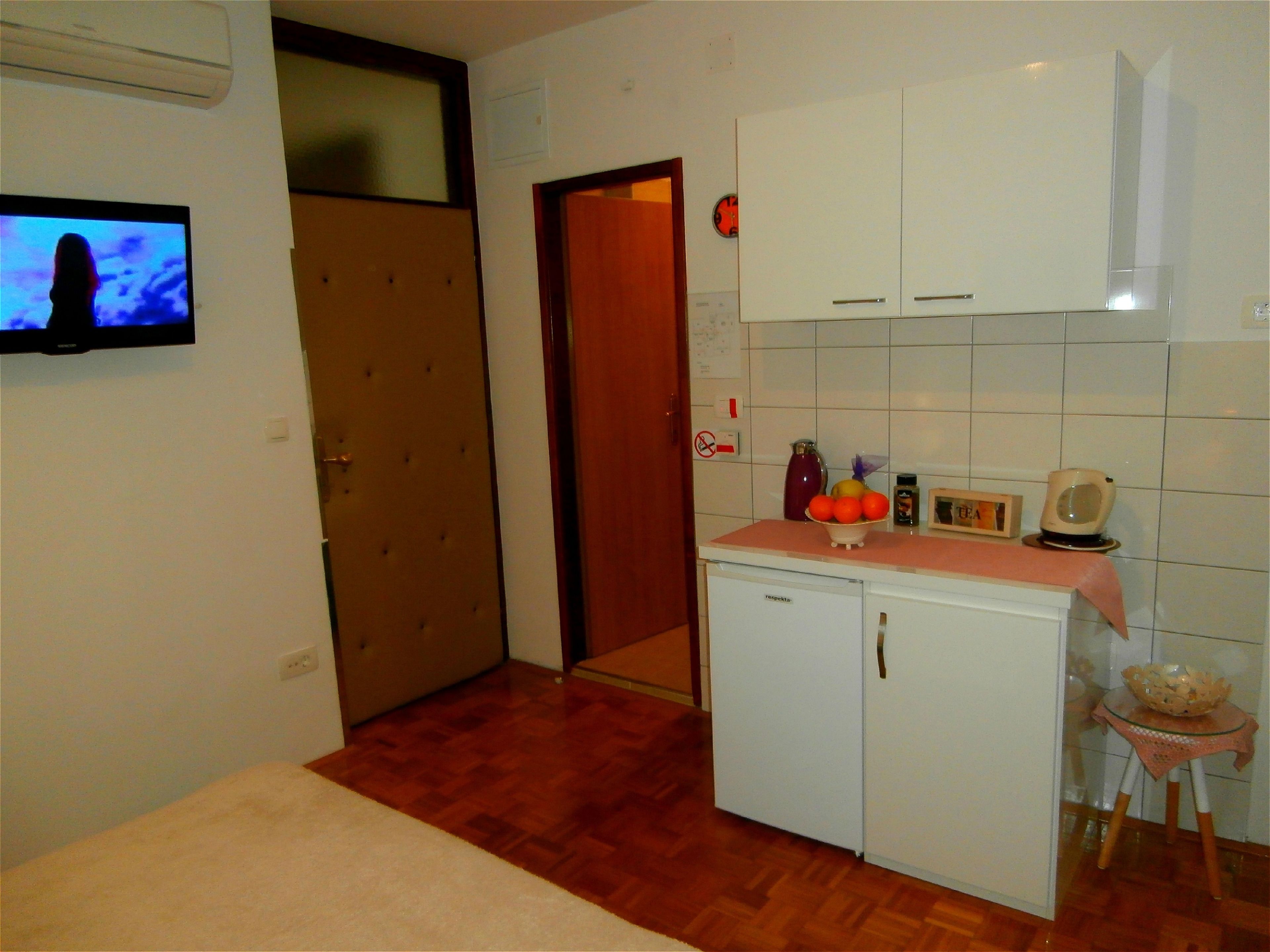 A compact kitchen with a refrigerator, a small sink, and a countertop with a fruit bowl and a kettle.