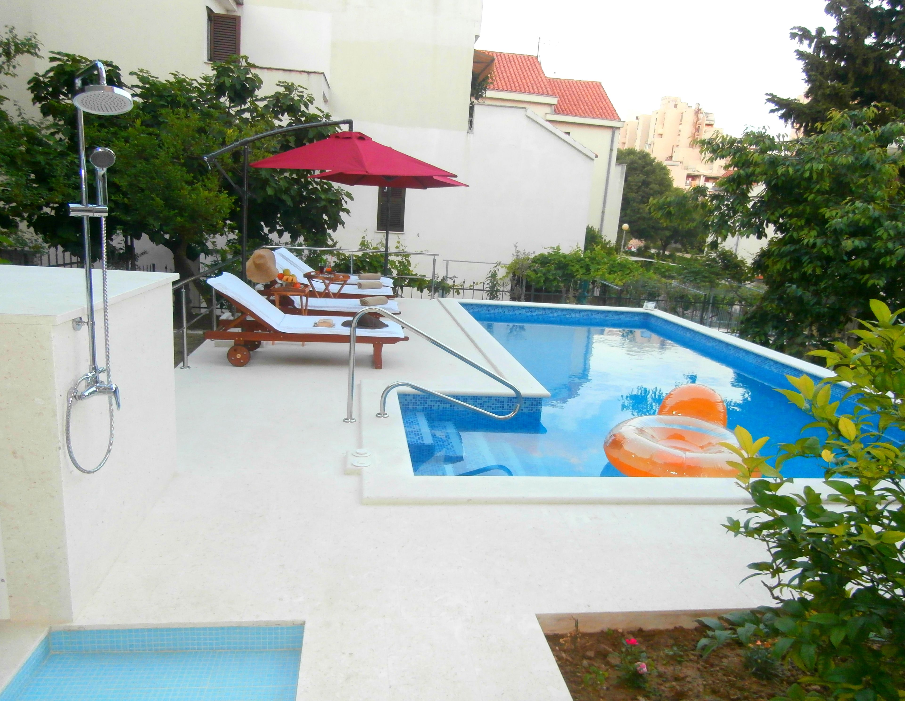 A swimming pool with two lounge chairs and a red umbrella, surrounded by greenery.