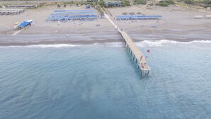 Eine Luftaufnahme eines Strandes mit einem Steg, der ins Meer führt, mehreren Reihen von Strandsonnenschirmen und einigen Personen auf dem Steg.