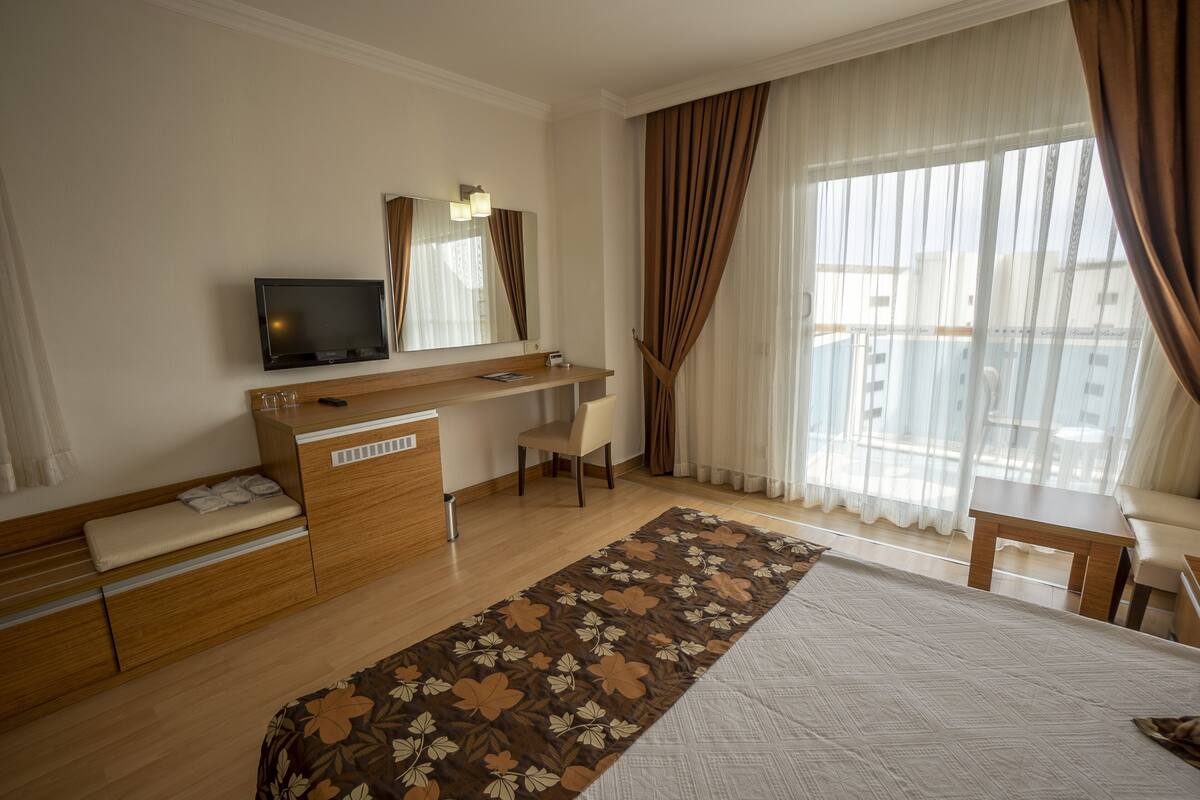 A hotel room with a bed, a desk with a television, a chair, a window with curtains, and a view of the city.