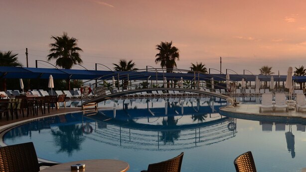 Ein Poolbereich mit Liegestühlen und Sonnenschirmen, umgeben von Palmen und einem Sonnenuntergang.