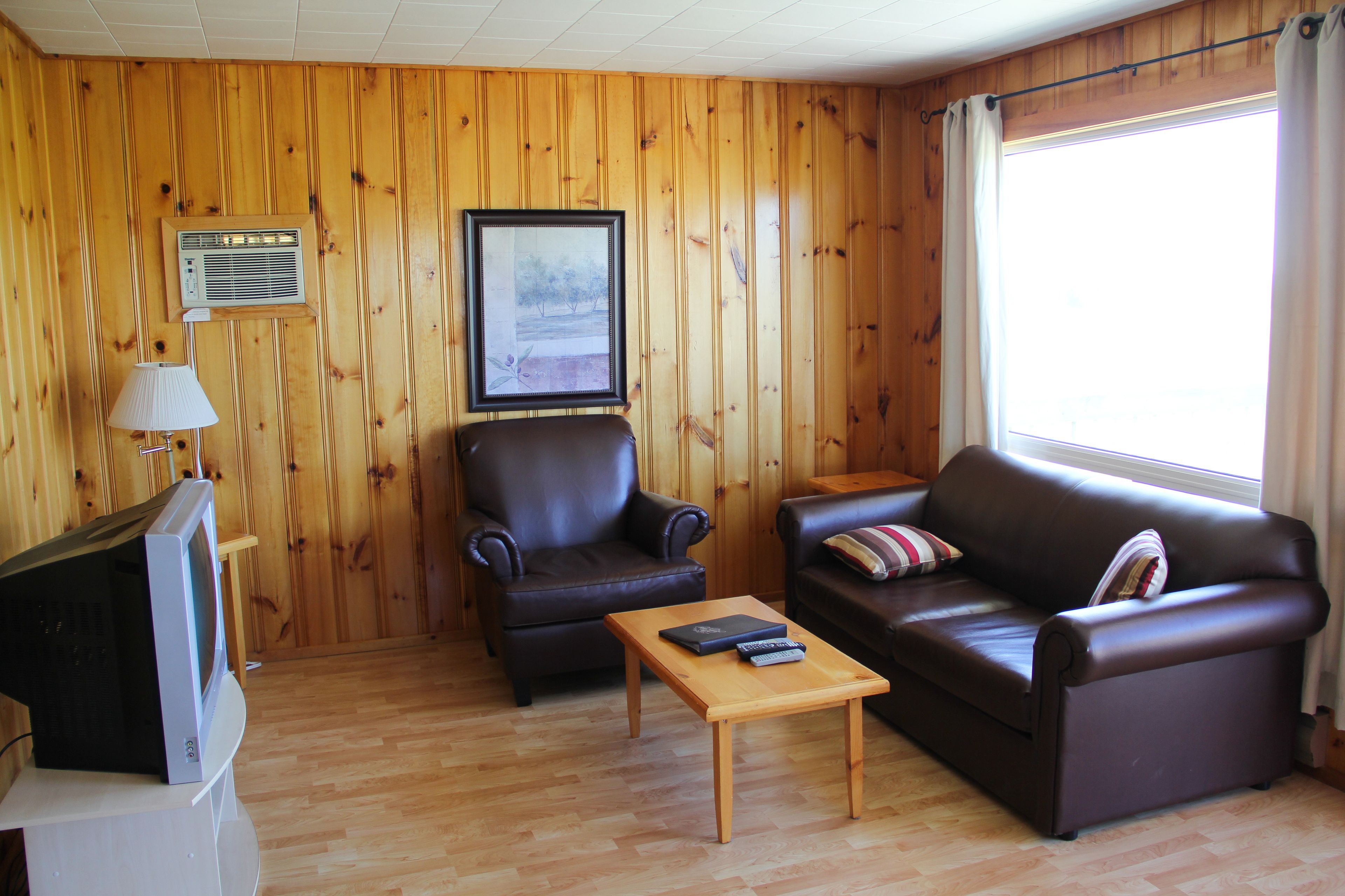 Una habitación con paneles de madera, un sofá de cuero, una mesa de centro de madera, un televisor sobre un mueble bajo y una ventana con cortinas.