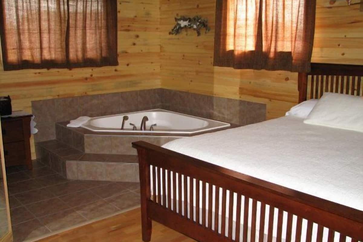 A wooden cabin bedroom with a large bed, a wooden headboard, a built-in bathtub, and a small bedside table.