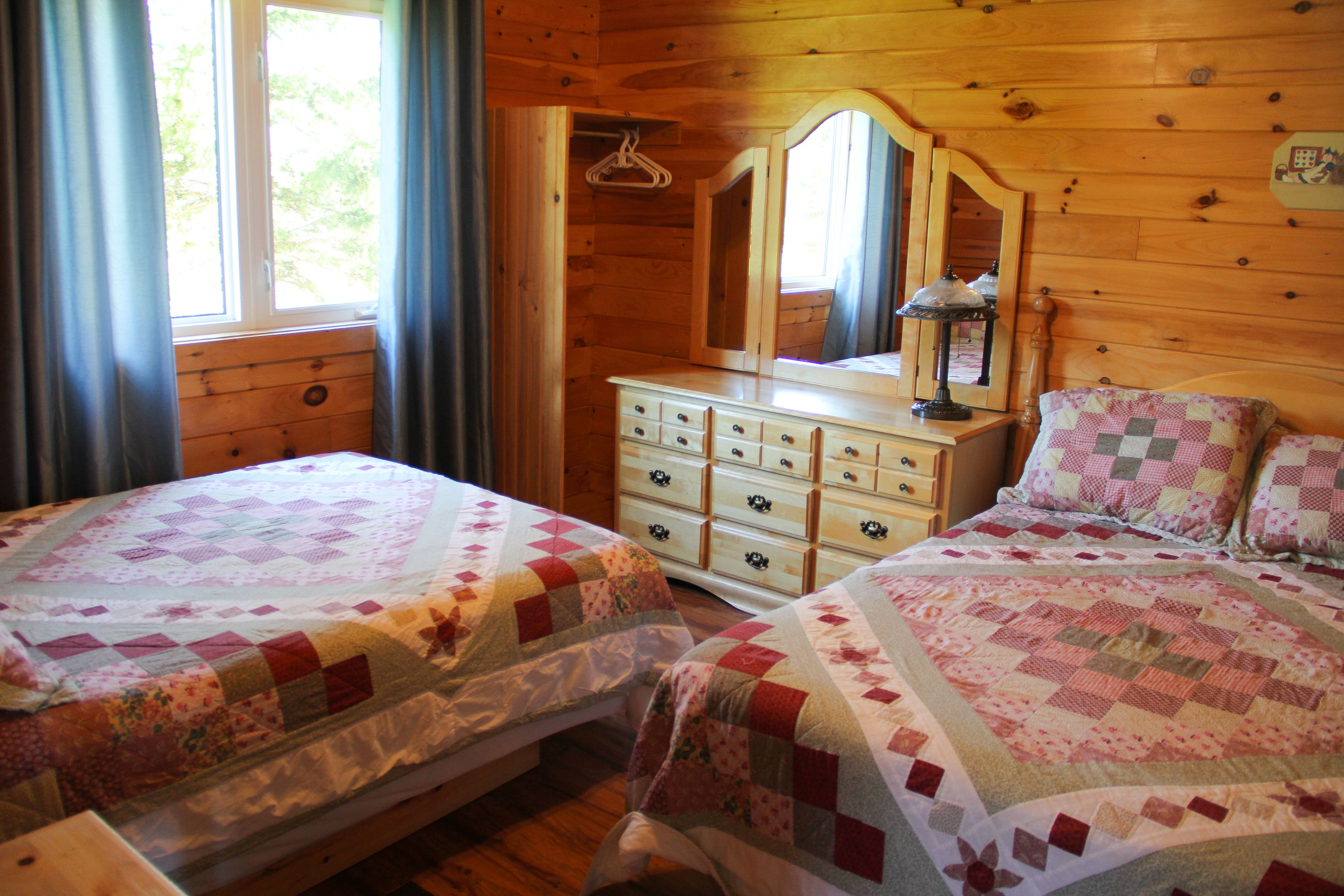 A wooden cabin bedroom with two beds, a dresser, a mirror, and a window with curtains.