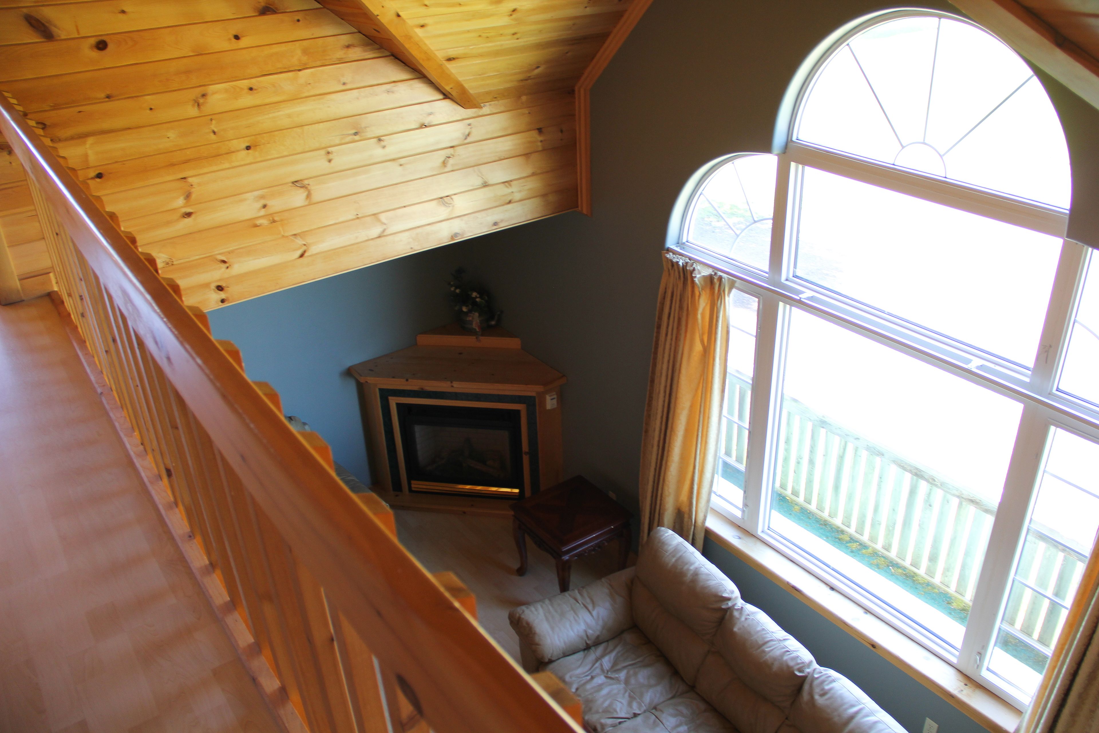 A wooden staircase leading to a lofted area with a fireplace, a cushioned seating area, and a large window with curtains.