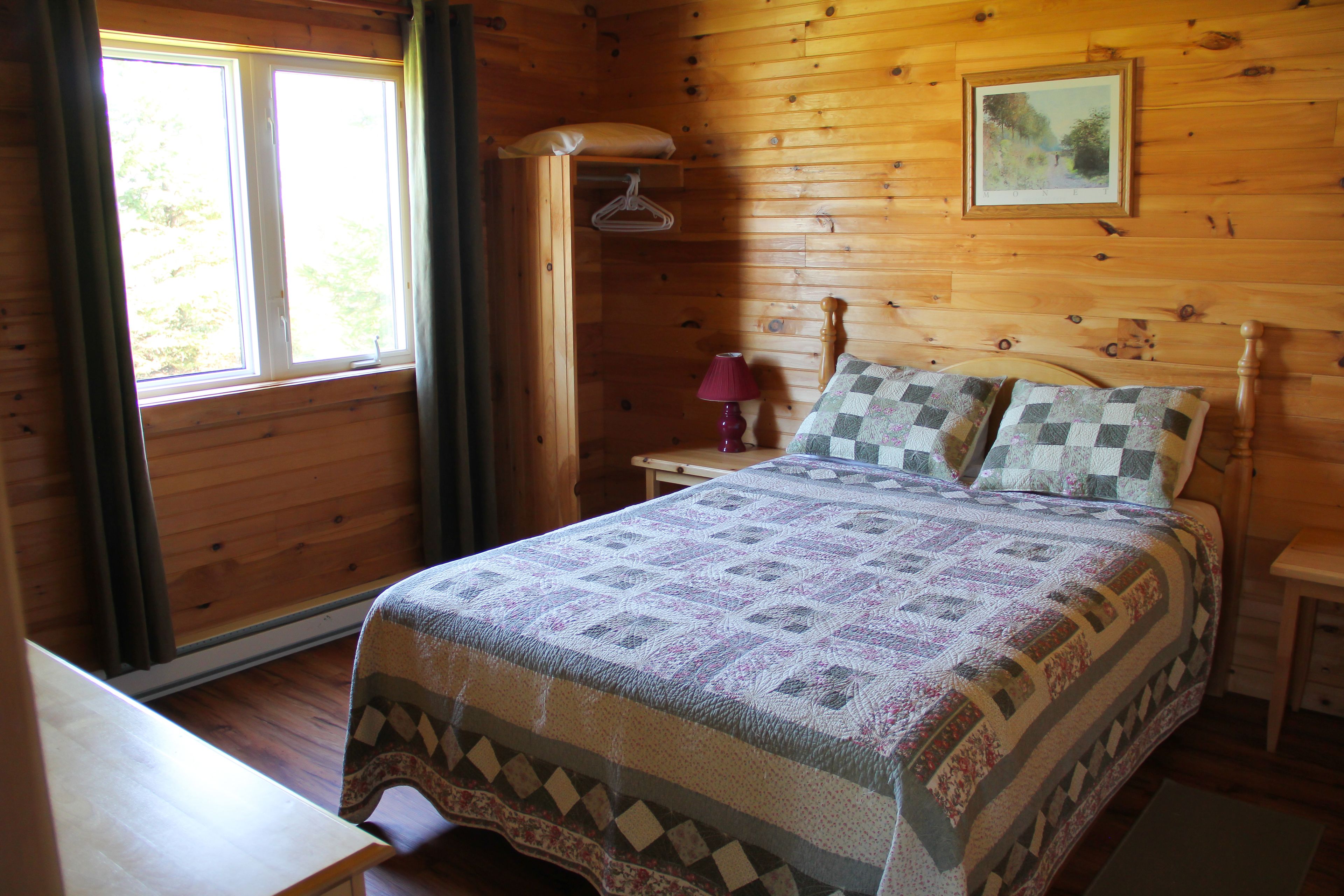Un dormitorio de cabaña de madera con cama, mesita de noche, lámpara y ventana con cortinas.