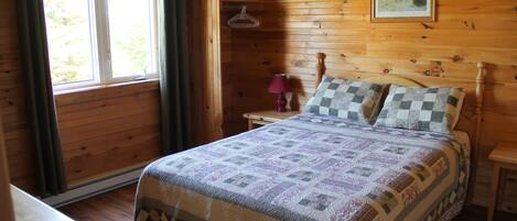 Un dormitorio de cabaña de madera con cama, mesita de noche, lámpara y ventana con cortinas.