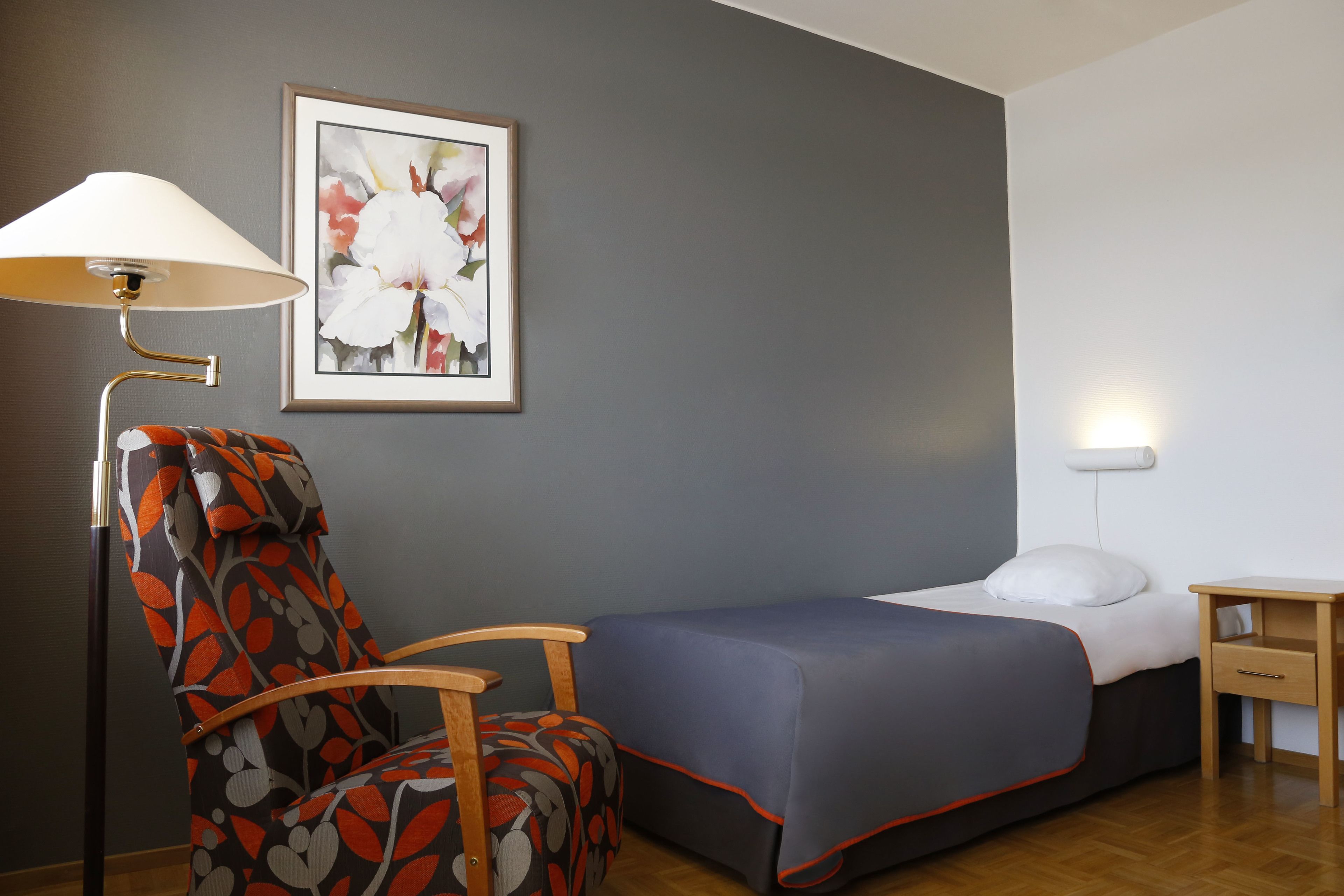 A bedroom with a single bed, a chair with a floral pattern, a lamp, and a framed picture on the wall.