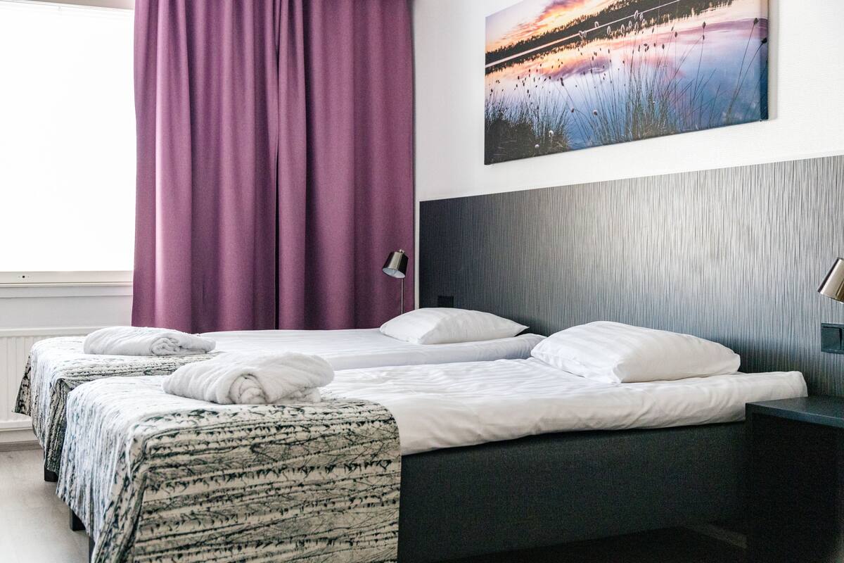 Una habitación de hotel con dos camas, un ventanal con cortinas moradas, una lámpara de pared y un cuadro de un atardecer sobre un lago.