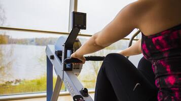 Uma pessoa usando uma bicicleta ergométrica com visor digital, sentada em frente a uma janela com vista para um corpo de água.
