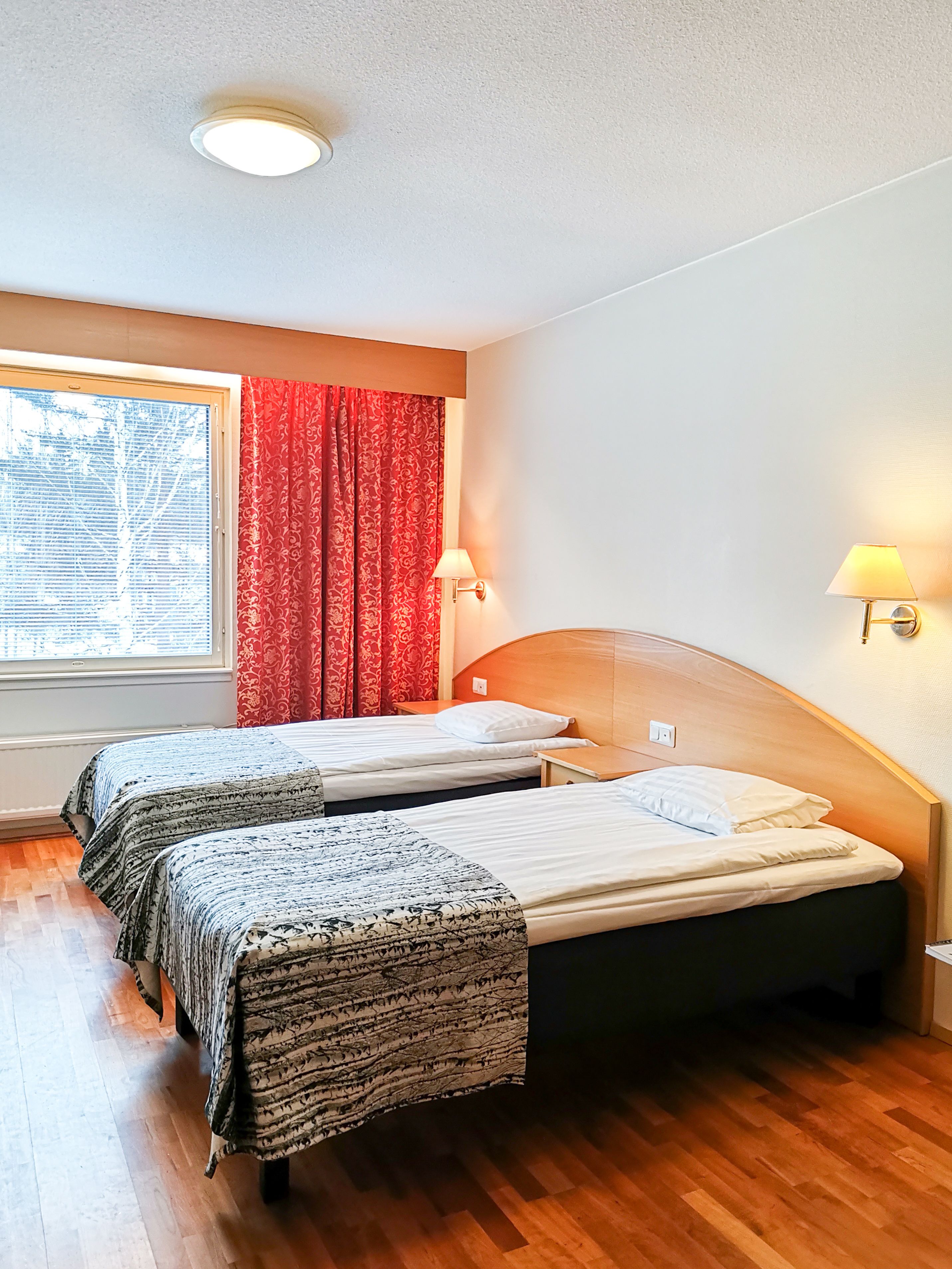 A room with two beds, a window with red curtains, a lamp, and a ceiling light.