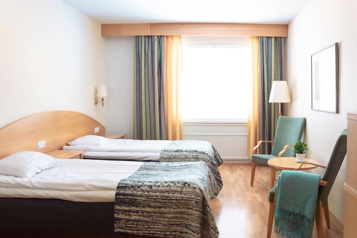 Habitación de hotel con dos camas, una silla, una mesita, una lámpara y una ventana con cortinas.