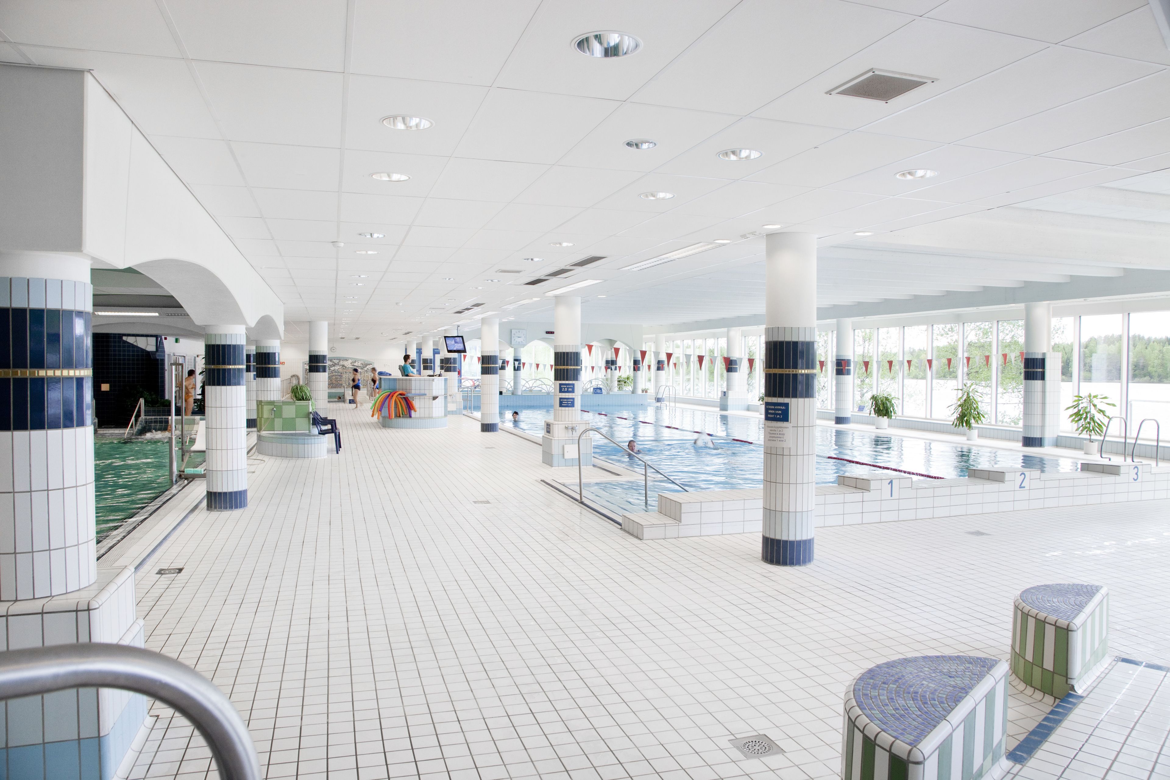 Un vaste espace intérieur abritant une piscine, avec des sols carrelés, plusieurs bassins et un comptoir d’accueil.