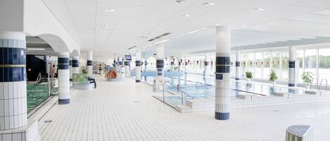 A spacious indoor swimming pool area with tiled floors, multiple pools, and a reception desk.