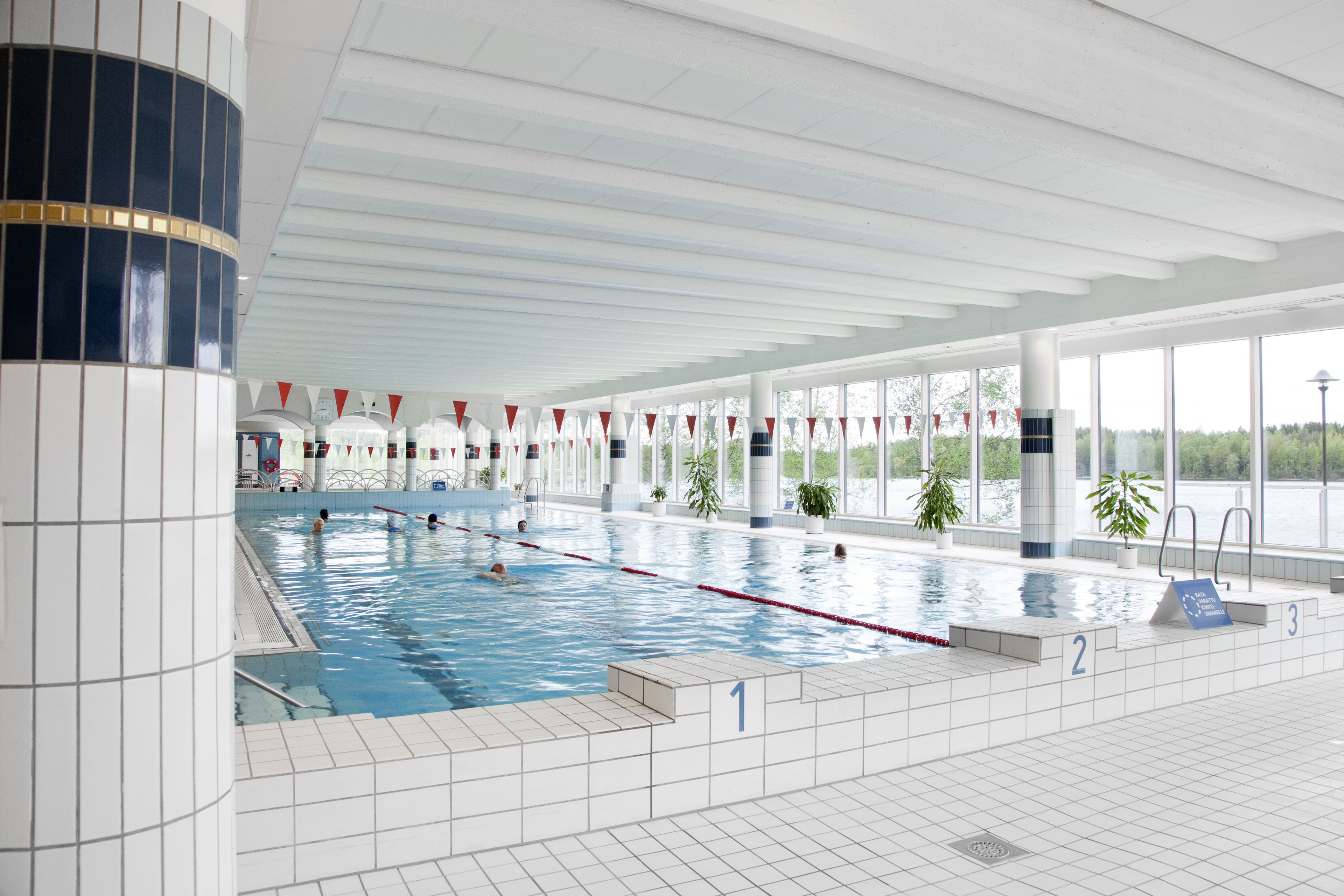 An indoor swimming pool with clear lanes, a tiled floor, and a view of the outdoors through large windows.