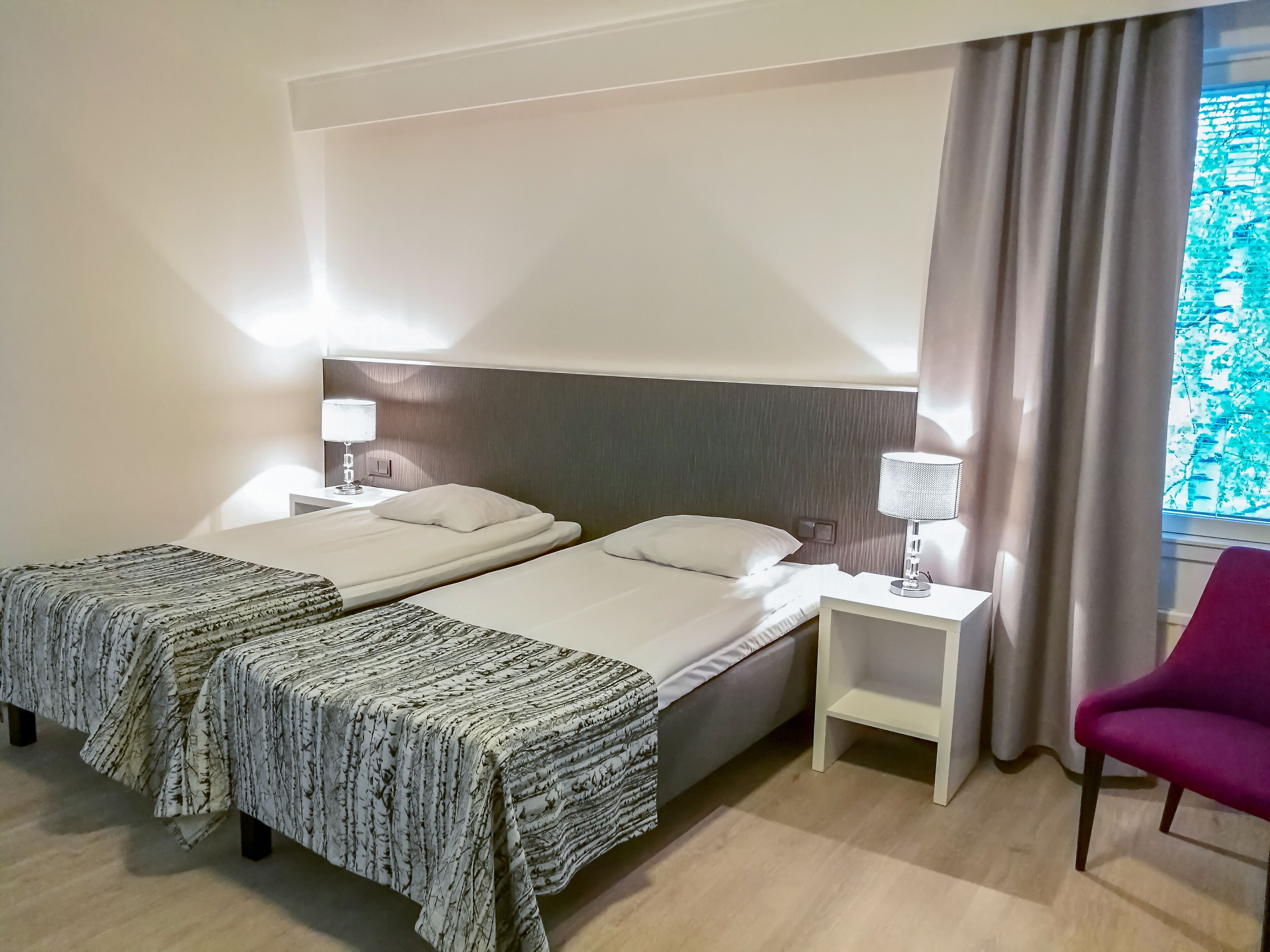 Een hotelkamer met twee bedden, een nachtkastje met een lamp, een stoel en een raam met gordijnen.