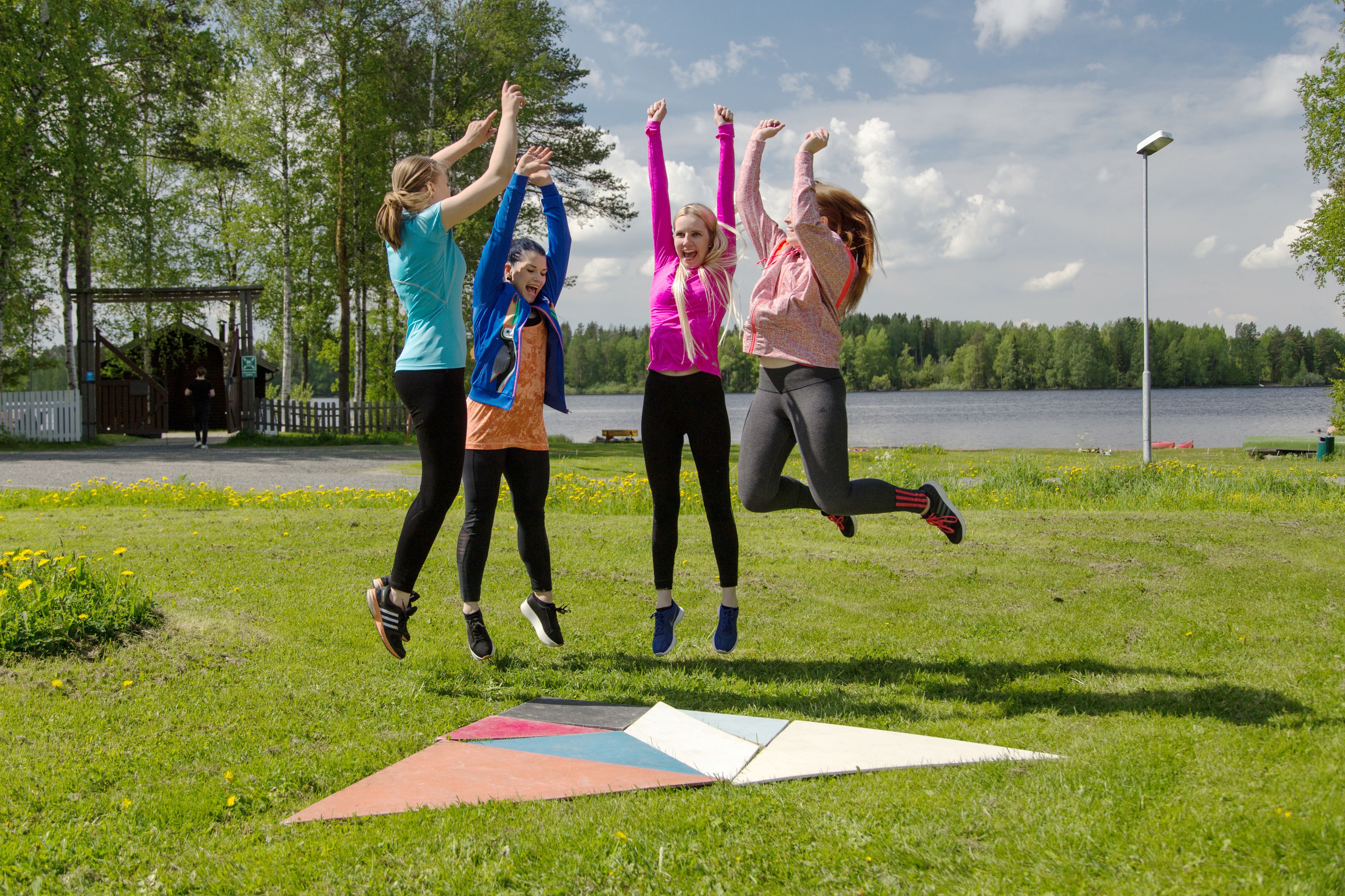 Ryhmä ihmisiä hyppää ruohikkoisella alueella, taustalla näkyy järvi.