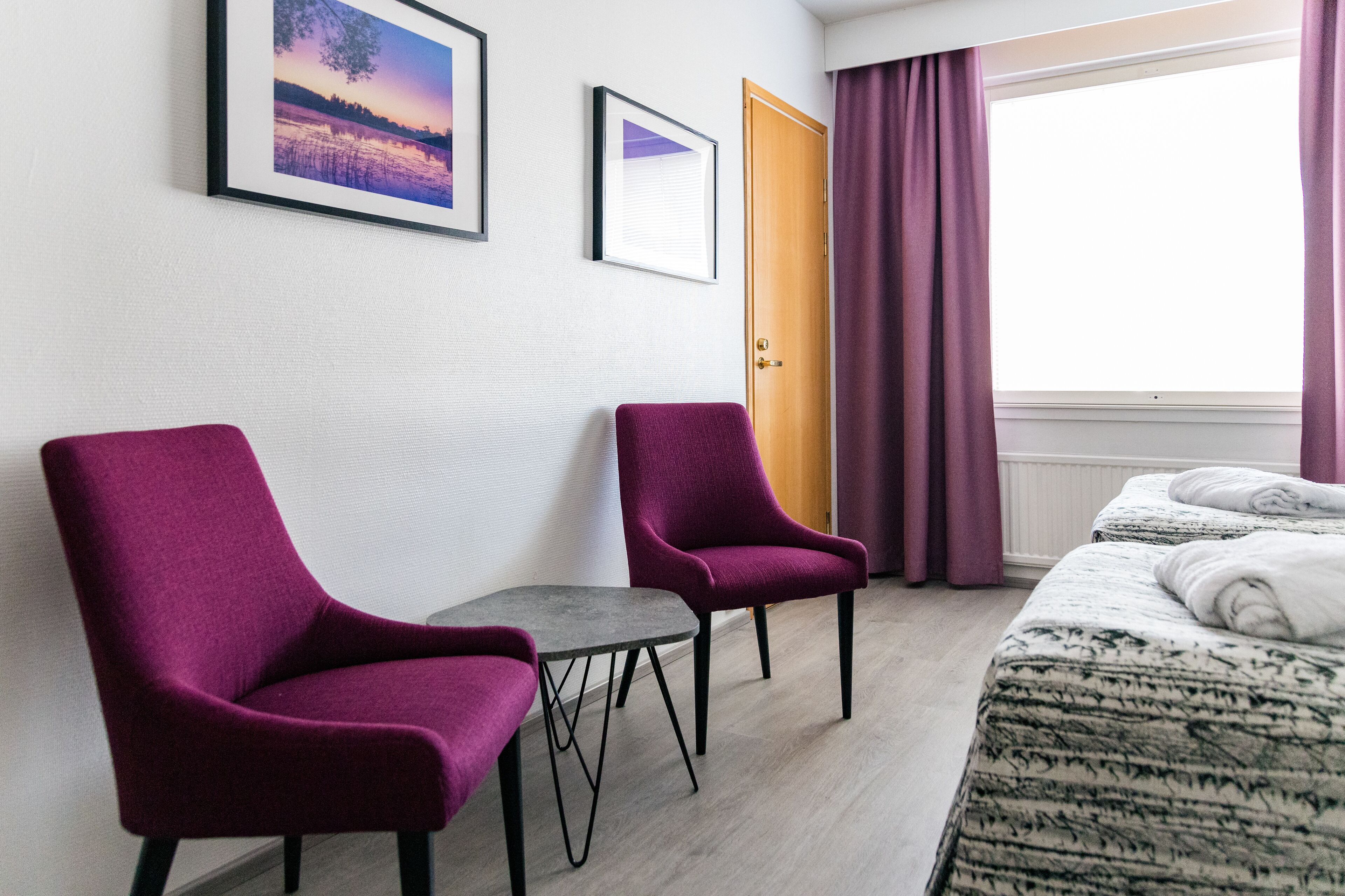 A hotel room with two purple armchairs, a small table, a bed with a patterned duvet, a window with purple curtains, and two framed pictures on the wall.