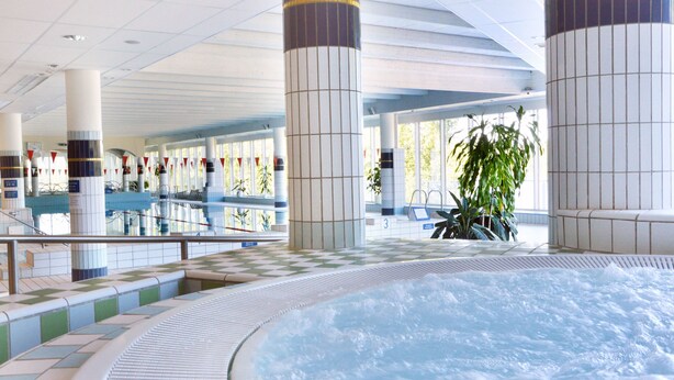 Amplia zona de piscina interior con jacuzzi, suelo de baldosas y grandes pilares.
