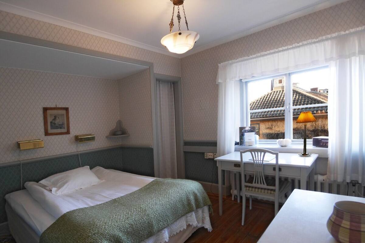 Hotelkamer met een bed, een bureau en een stoel. Er is een raam met gordijnen, een lamp op het bureau en een schilderij aan de muur.