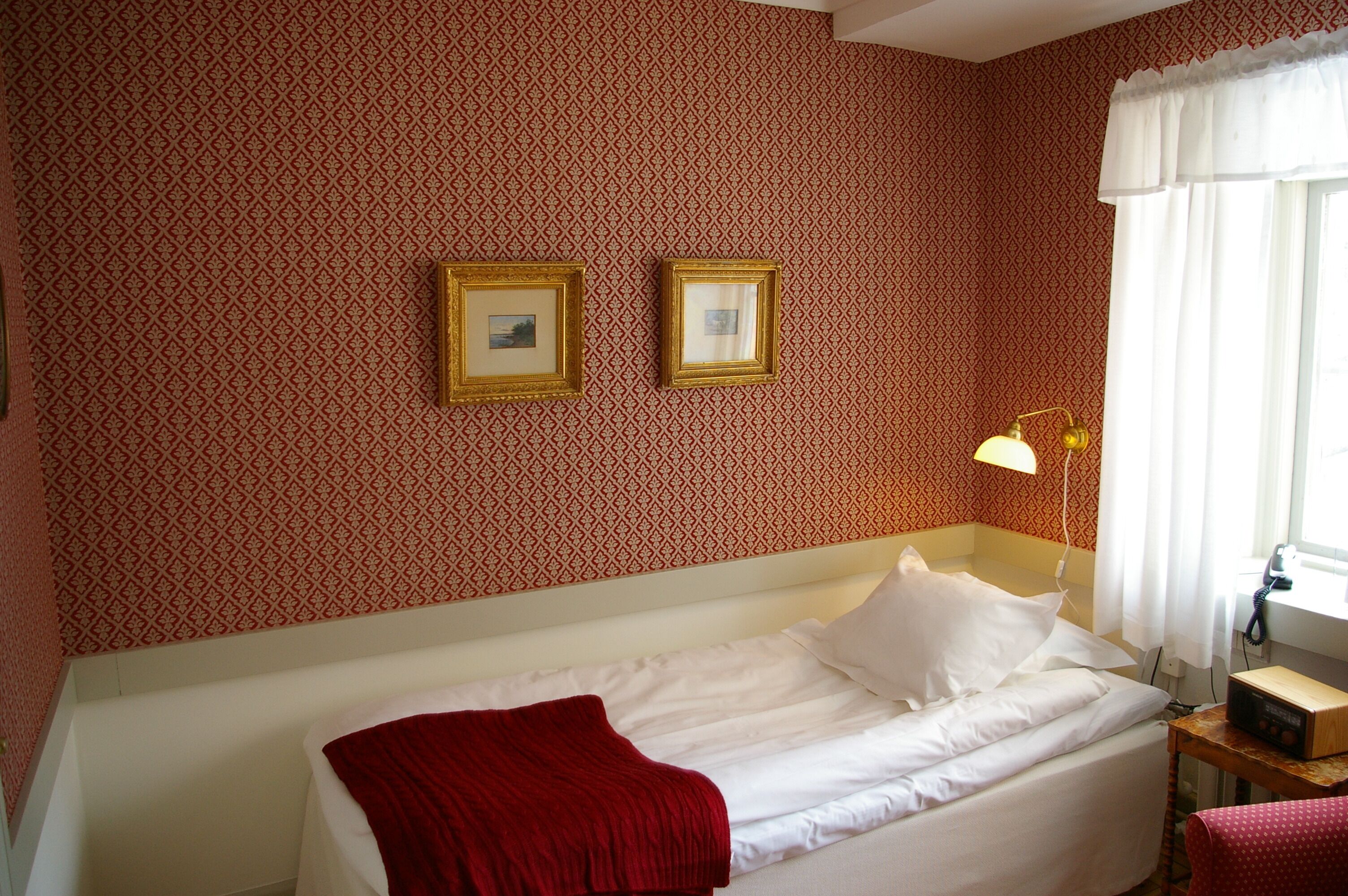 Una cama individual con ropa blanca, una manta roja y dos cuadros en la pared.
