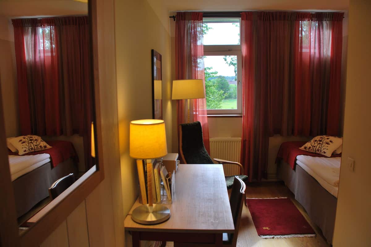 Een hotelkamer met twee bedden, een bureau met een lamp, een stoel en uitzicht op groen.