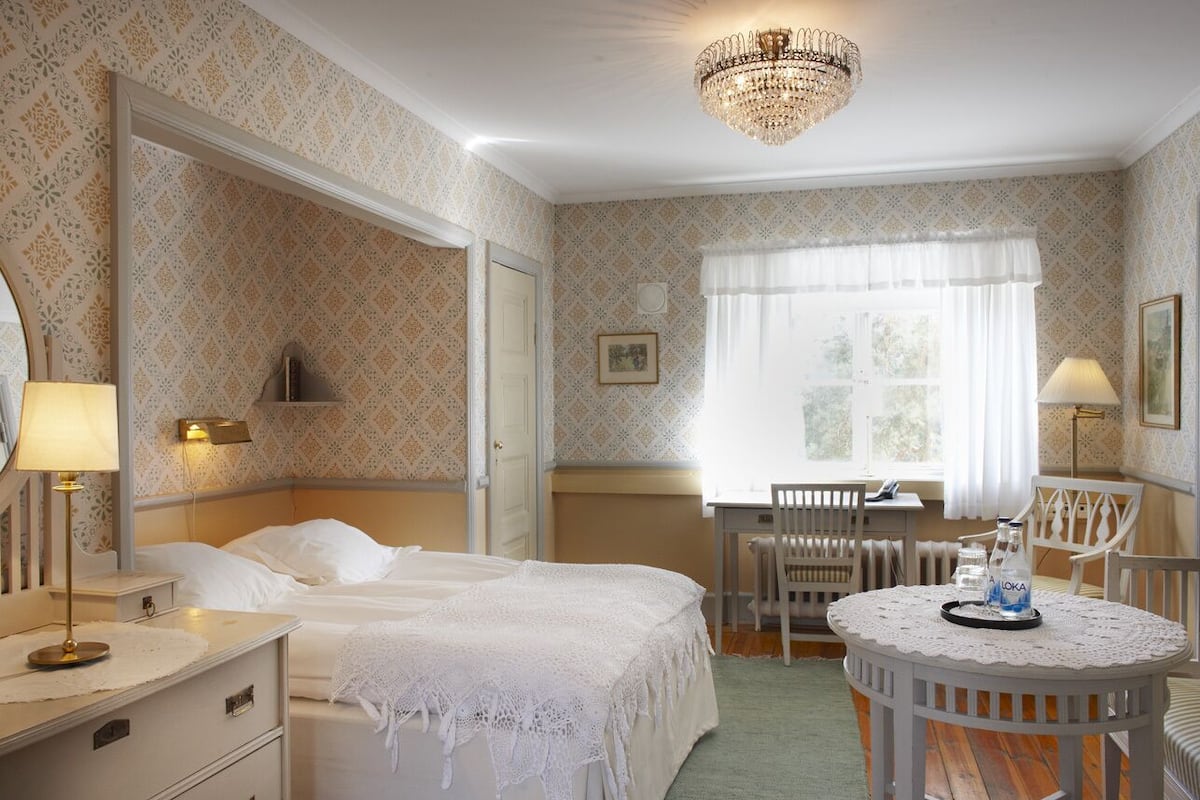 Een hotelkamer met een bed, nachtkastje, lamp, kroonluchter en een eethoek met een tafel en stoelen.