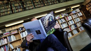 Une personne est assise dans une bibliothèque, en train de lire un livre, avec des étagères remplies de livres en arrière-plan.