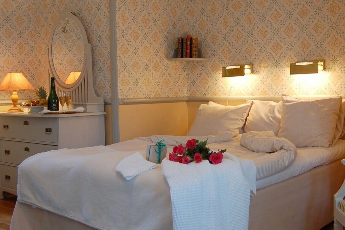 Een net opgemaakt bed met een witte dekbedovertrek en kussens, een nachtkastje met een lamp en een fles, een stoel en een muur met behang met een patroon en een plank met boeken.