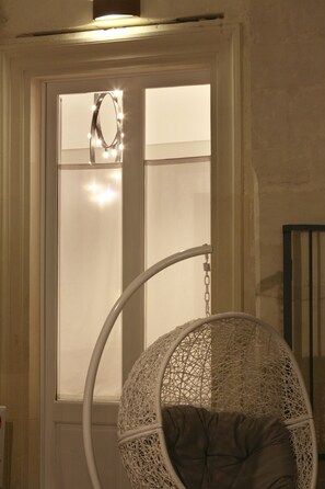 A room with a white wicker hanging chair with a cushion, a white-framed window with frosted glass, and a light fixture above the window. - Santa Marta Suites & Apartments (Lecce)