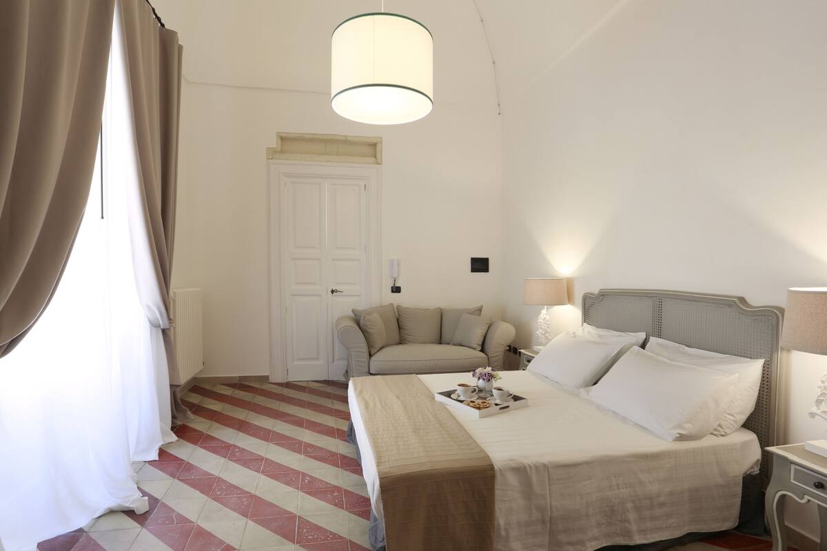 A hotel room with a bed, a sofa, a lamp, and a window with curtains.