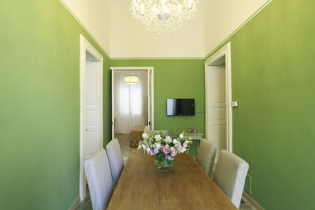 A dining area with a wooden table, chairs, a vase of flowers, a chandelier, and a television on the wall.