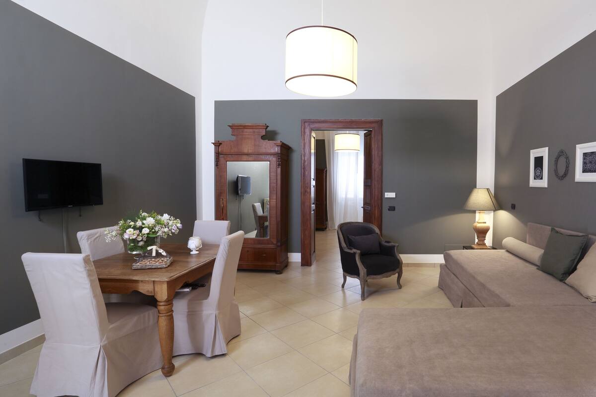 A living room with a sofa, a dining table with chairs, a TV mounted on the wall, and a lamp.