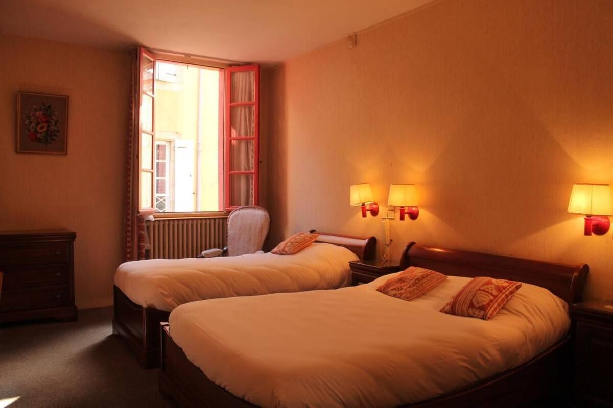 A hotel room with two beds, a window with curtains, a radiator, a chair, and wall-mounted lamps.