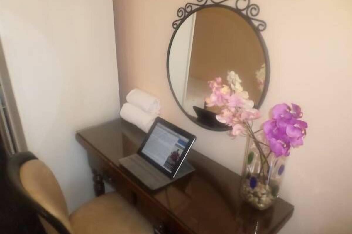 A small desk with a laptop, a mirror, a vase with flowers, and a chair.