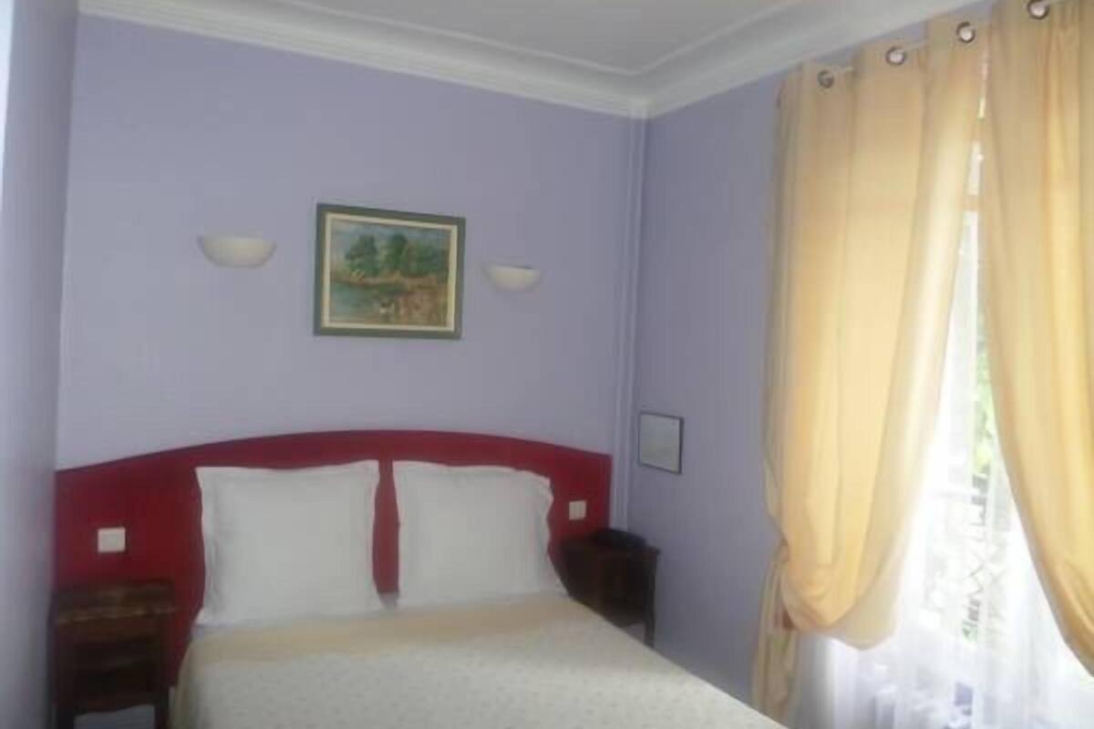 A bedroom with a bed, a nightstand, a wall-mounted lamp, and a window with curtains.
