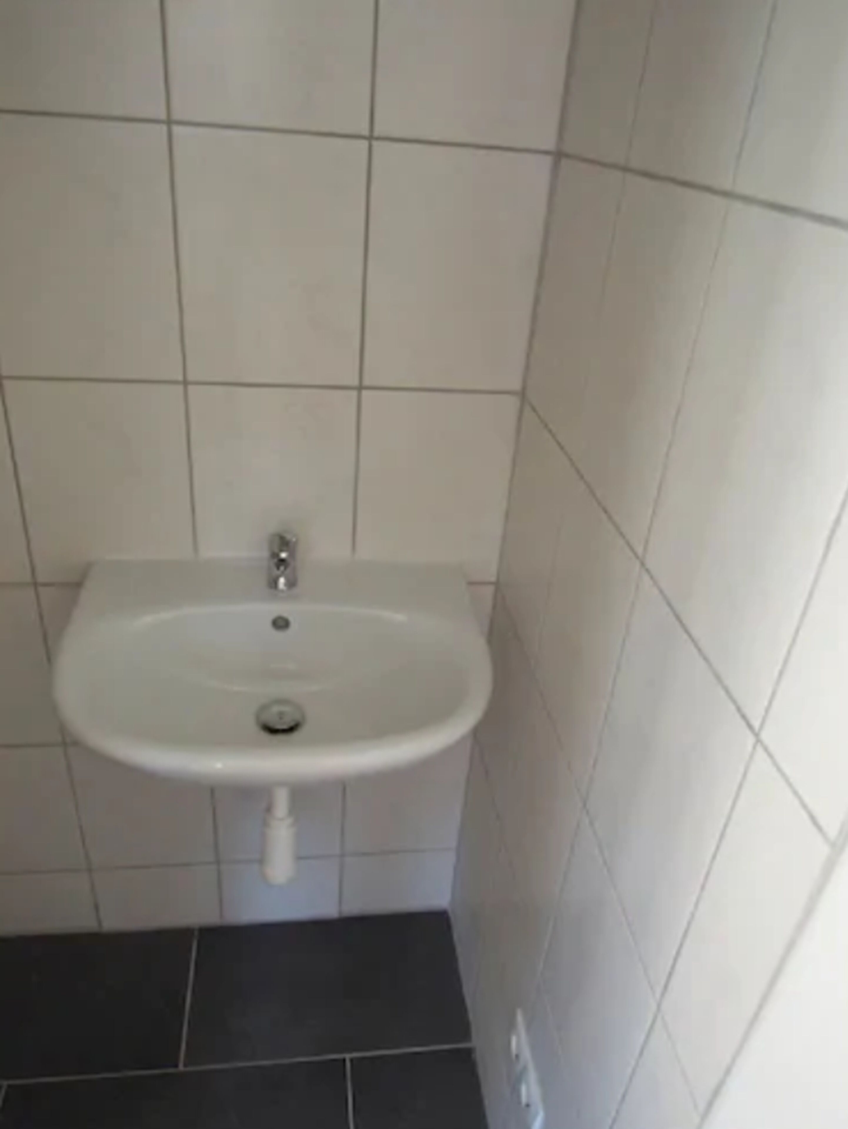 Ein kleines, weißes Waschbecken mit einem einzelnen Wasserhahn, vor einer gefliesten Wand.