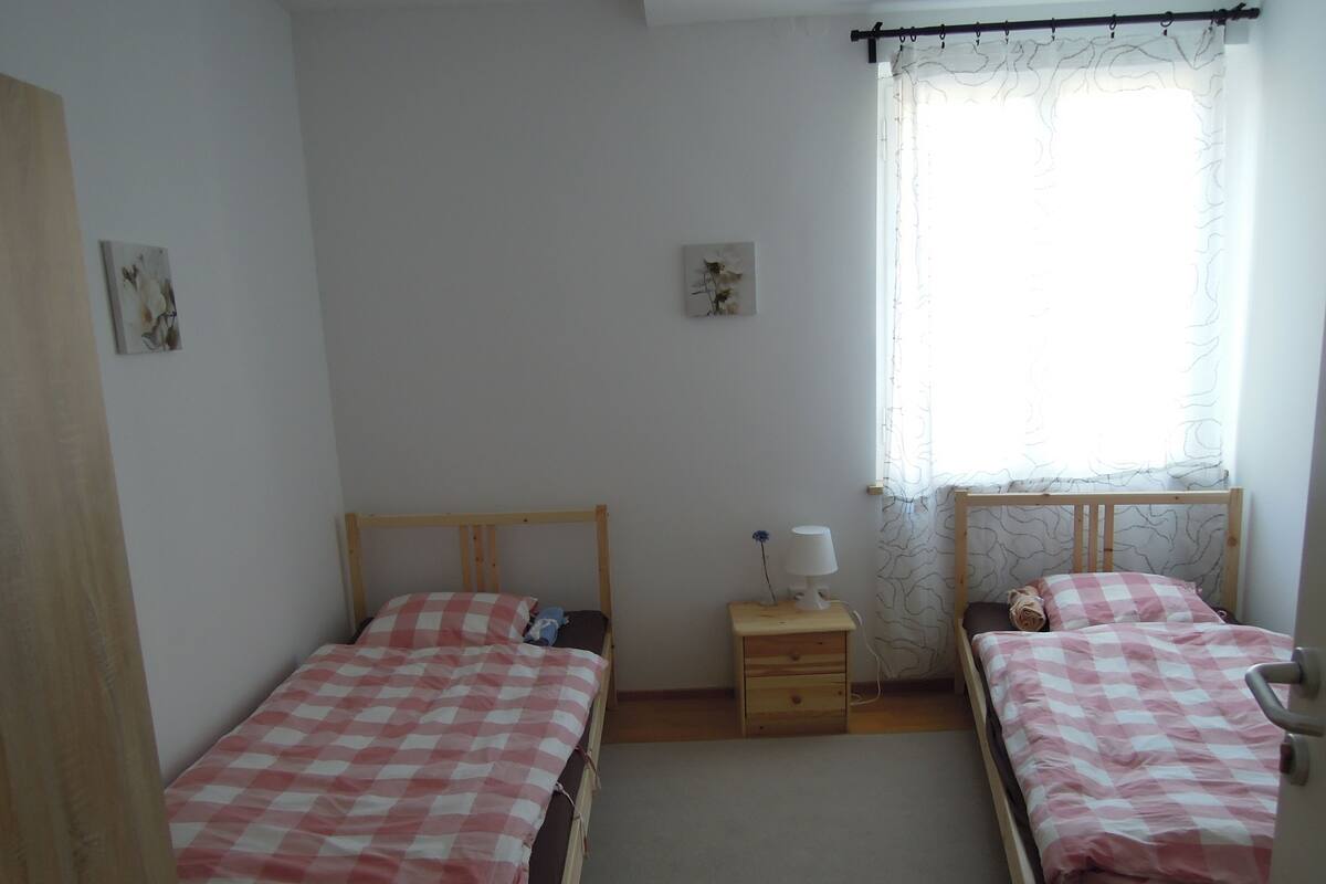 A small, clean room with two single beds, a nightstand with a lamp, and a window with sheer curtains.