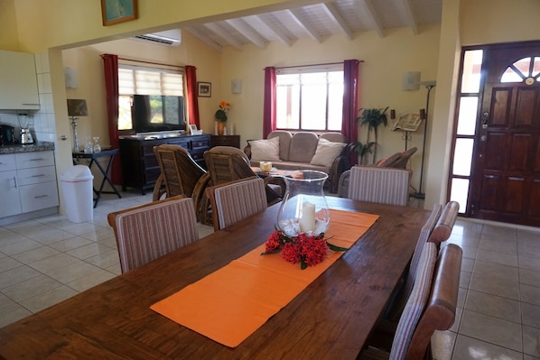 Un coin repas avec une table en bois, des sets de table orange et une pièce maîtresse décorative. On y trouve des chaises autour de la table, un canapé et un téléviseur à l’arrière-plan.