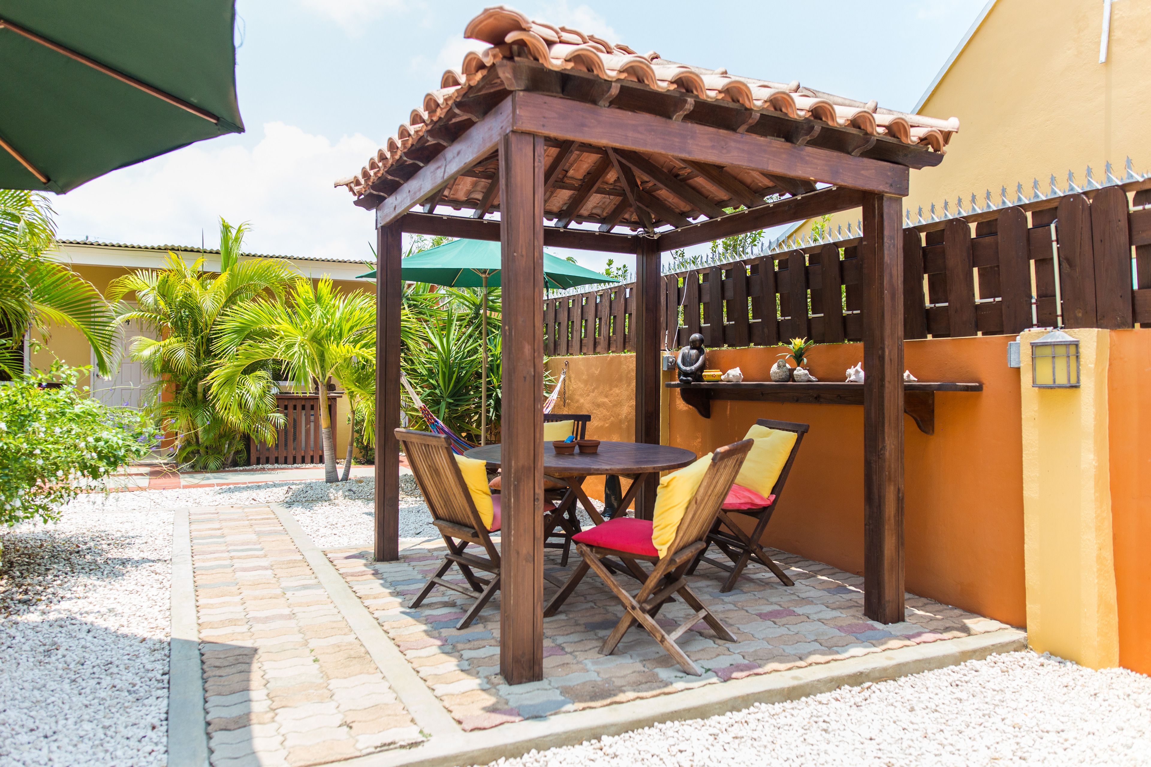 Une pergola en bois avec un toit en tuiles, une table ronde entourée de chaises et un coin bar avec un mur jaune et une clôture en bois.