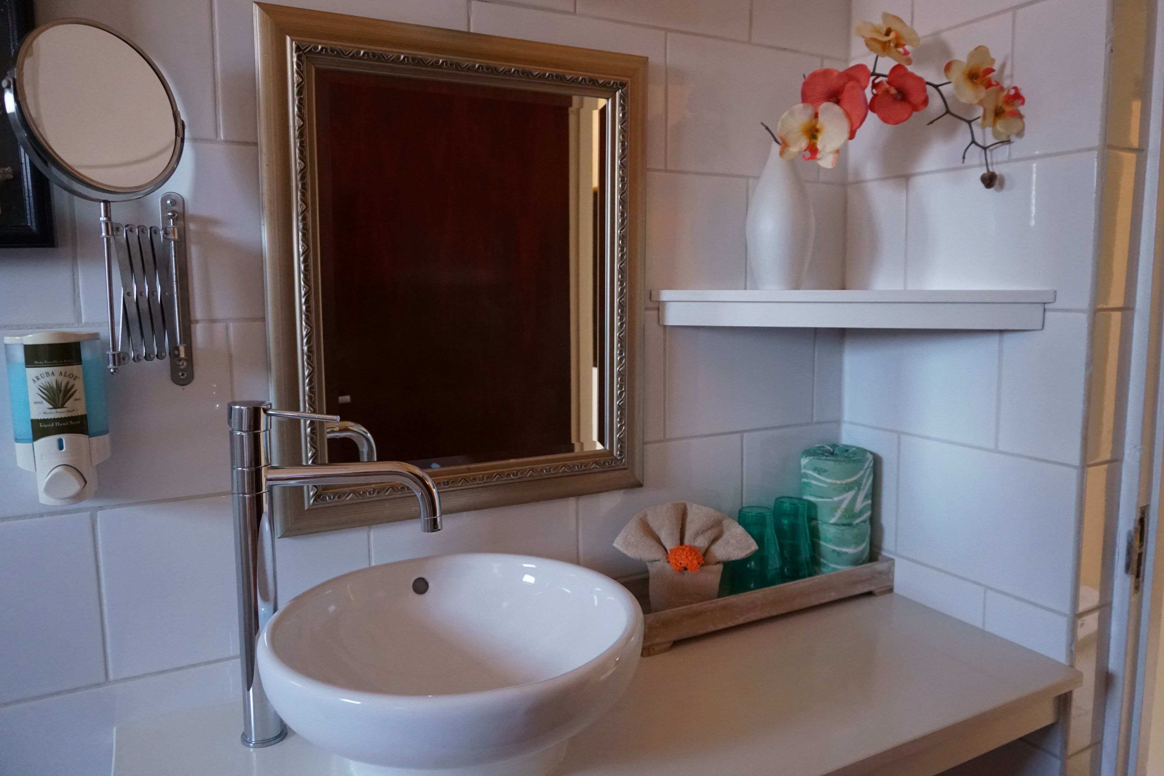 Een badkamer met een wit wastafelmeubel, een spiegel met gouden lijst, en een plankje met decoratieve bloemen en toiletartikelen.