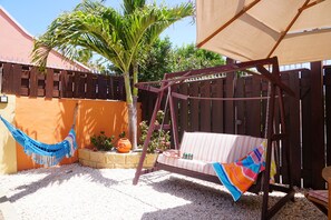 Un hamac, une balançoire et un banc rembourré dans un coin jardin agrémenté d’une clôture en bois et d’un palmier.