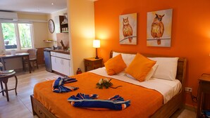 Habitación de hotel con una cama grande, ropa de cama naranja, un escritorio con silla, una pequeña zona de cocina y dos cuadros de búhos en la pared.