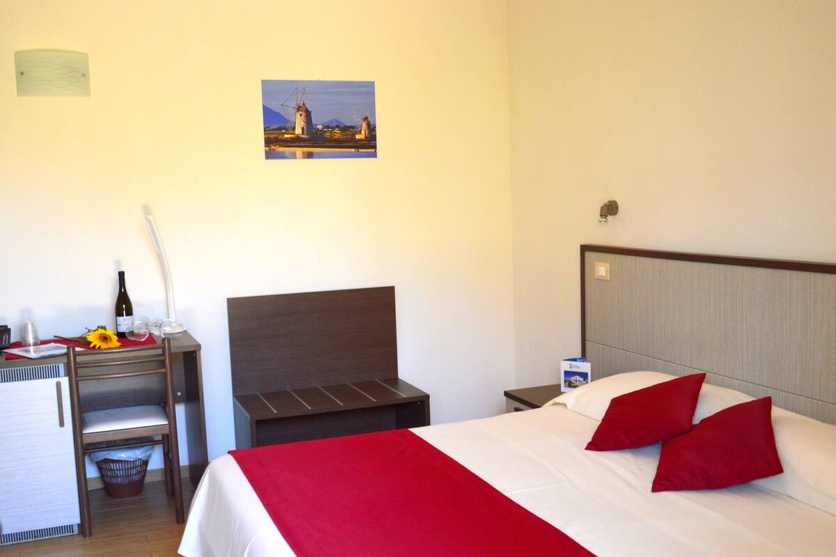 A hotel room with a bed, a desk, a chair, a small table with a wine bottle and glasses, a picture on the wall, and a window.