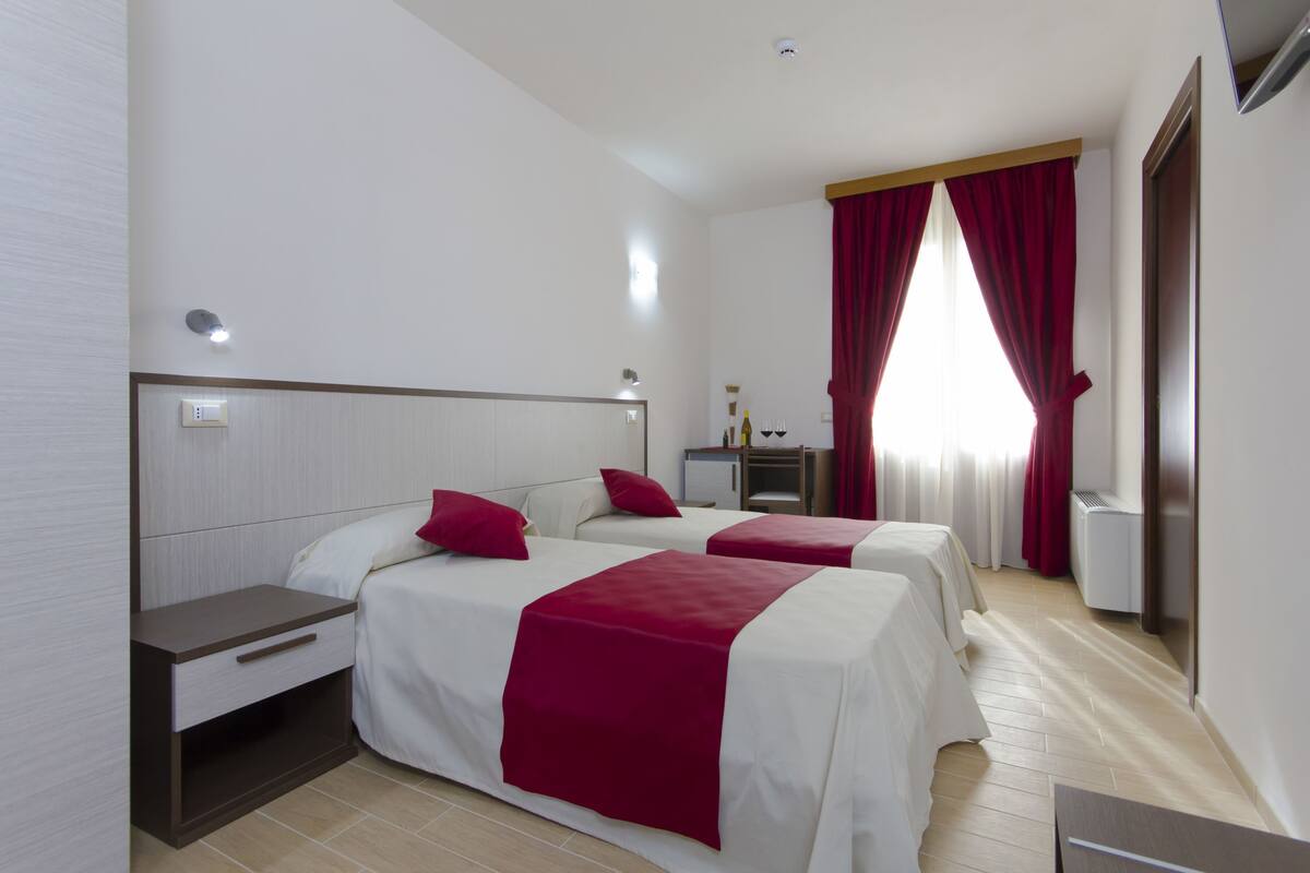 A hotel room with two beds, a desk, a small table, and a window with red curtains.