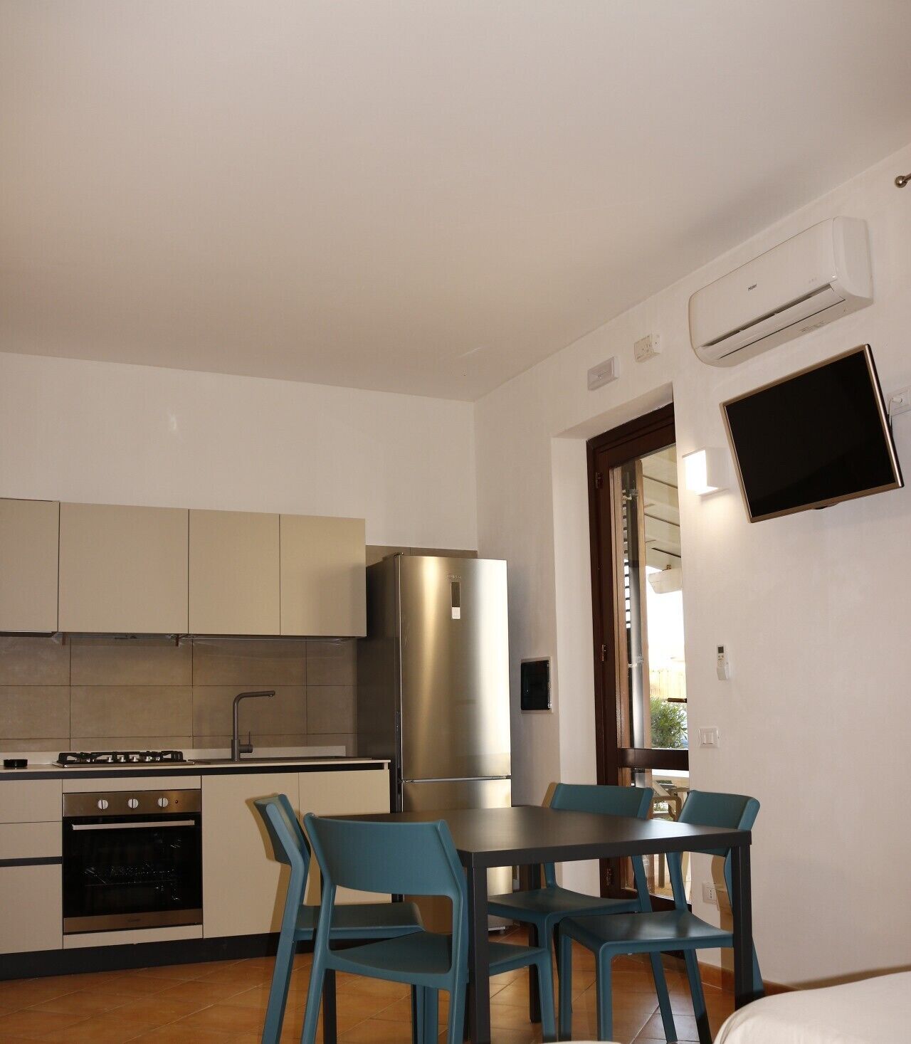 Een kleine keuken met moderne apparaten, een eethoek met blauwe stoelen, en een flatscreen televisie aan de muur.