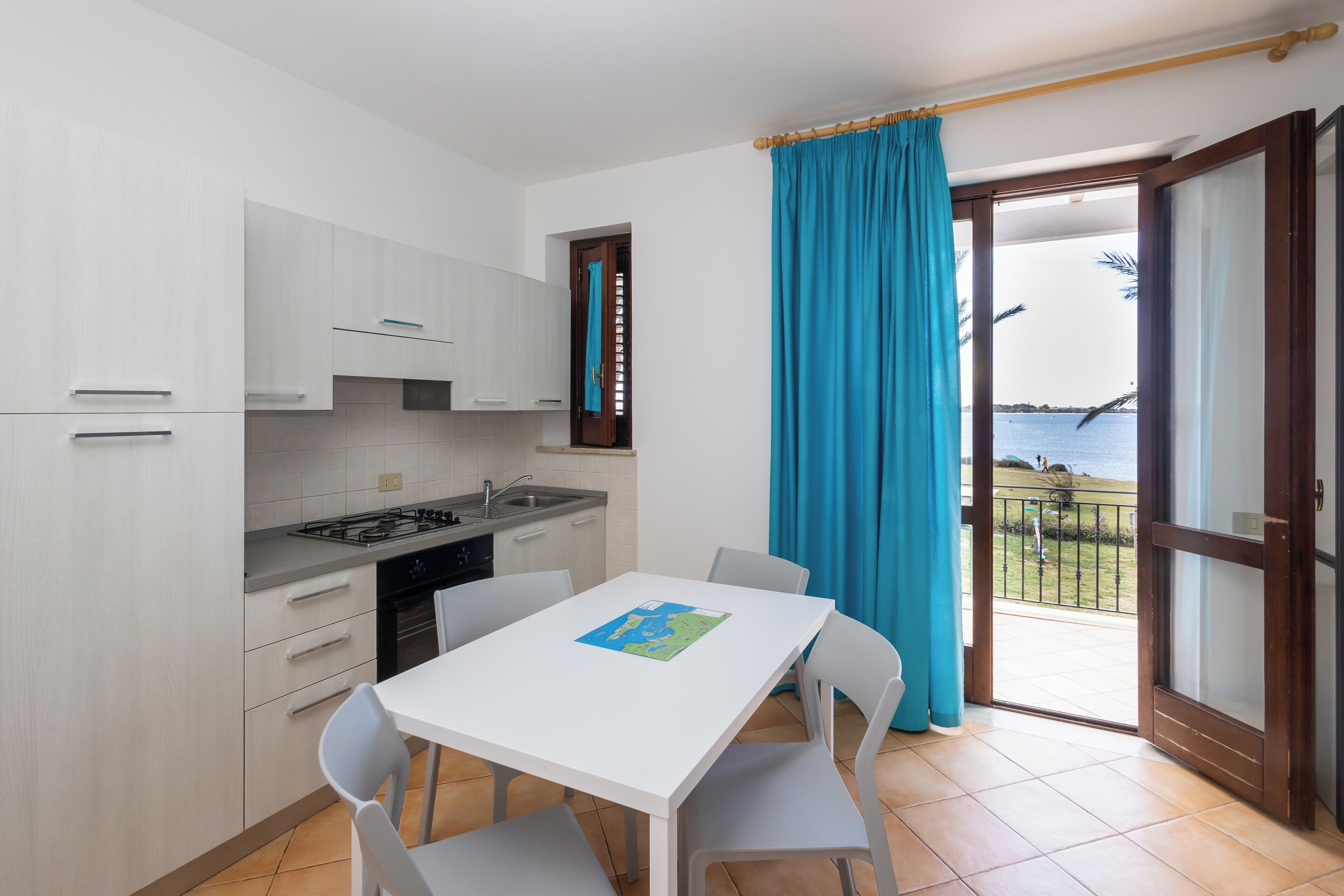 A modern kitchen with white cabinets, a gas stove, and a dining table with chairs. A window with a blue curtain provides a view of the sea.